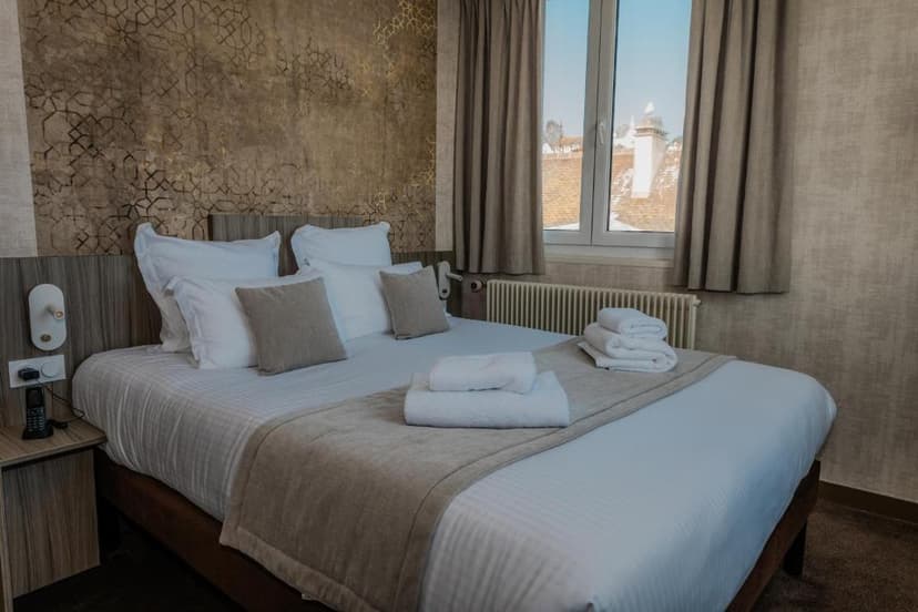 Hotel room with large bed, white linens, and patterned accent wall, view of rooftops outside window.