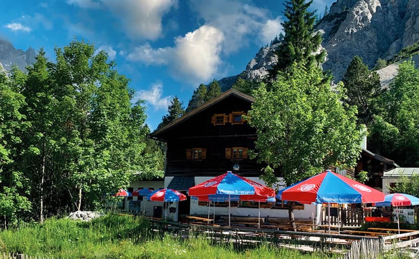Wimbachgrieshütte mountain hut with outdoor seating under red and blue umbrellas, surrounded by green trees and rocky peaks.