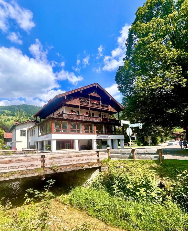 Hotel Königslinde building over a small stream with lush greenery and mountains in background.
