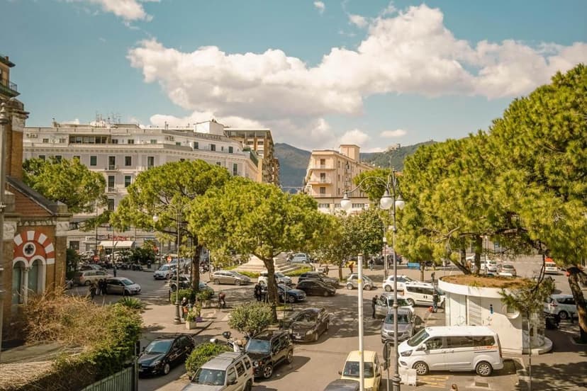 Busy city square with parked cars, tall pine trees, and multi-story buildings under a cloudy sky.
