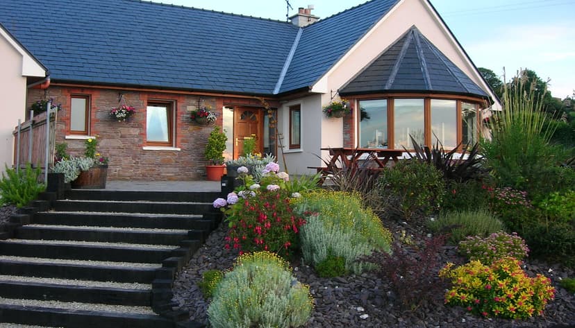 Stone house with dark roof, wooden steps, and lush garden with colorful flowers.