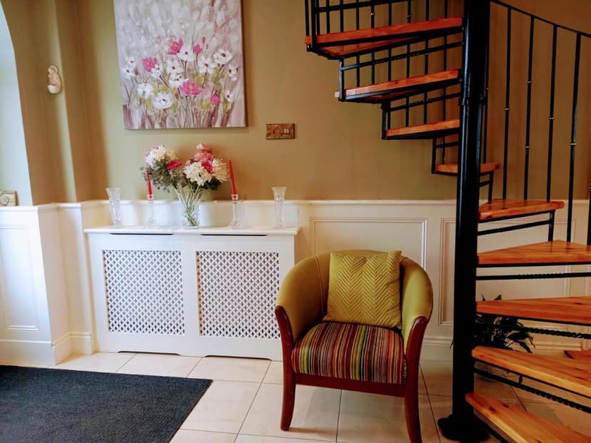 Spiral staircase next to an armchair, floral painting, and radiator cover in a room.