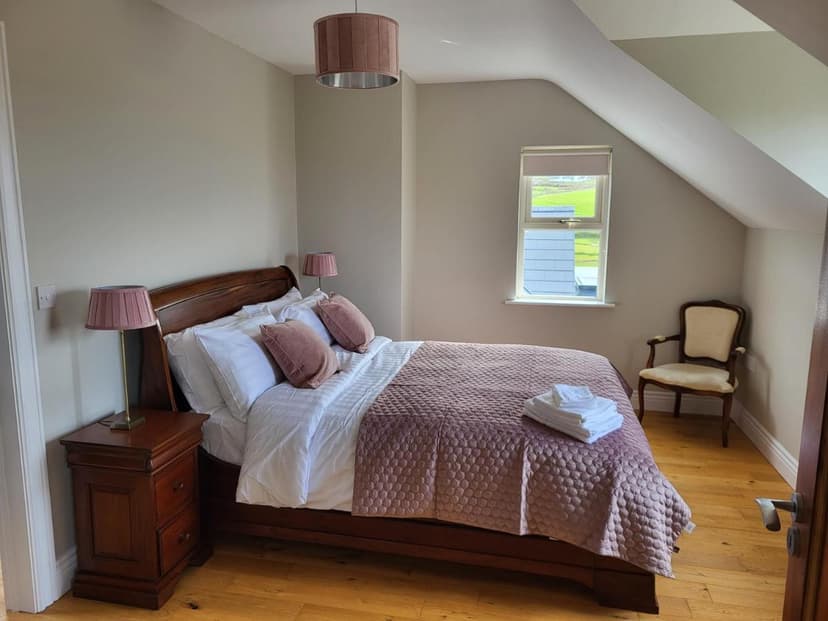 Bedroom with wooden bed, pink quilt, and attic sloped ceiling, view of green field from window.