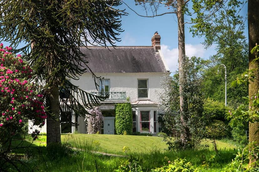 White lodge with slate roof framed by lush green trees and pink rhododendron blossoms.