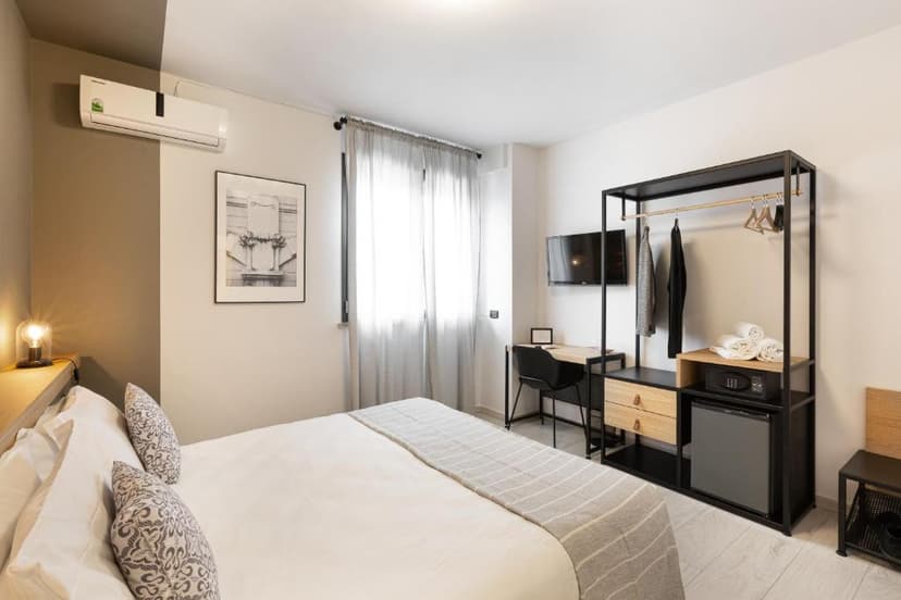Modern hotel bedroom with white bedding, open wardrobe, desk, and sheer window curtains.