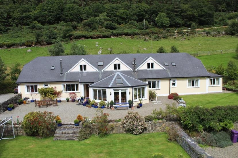 Large cream house with dark roof and conservatory set against a steep, wooded green hillside.