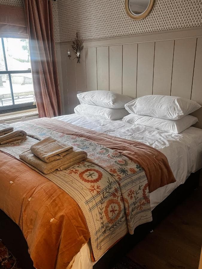 Bedroom with large bed, white pillows, embroidered throw, and window with brown curtains.