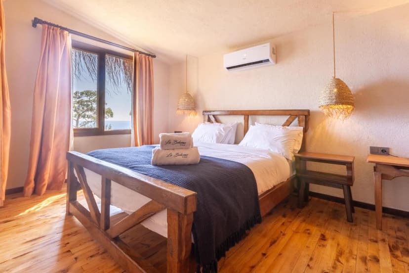 Bedroom with wooden bed, white linens, and view of sea and tree through window