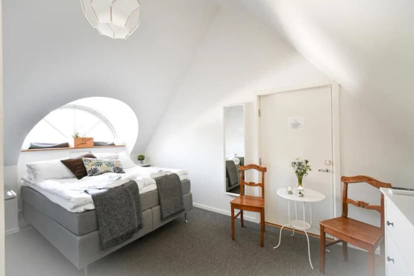 Attic hotel room with white walls, gray bed, and arched window at Kåseberga Gårdshotell.