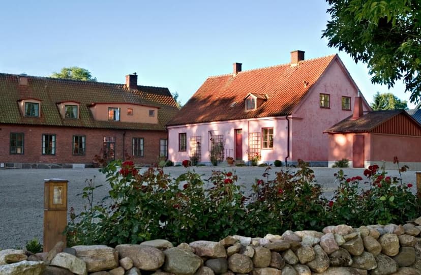 Two historic buildings with red tile roofs, one pink, in front of a gravel yard with roses and a stone wall.