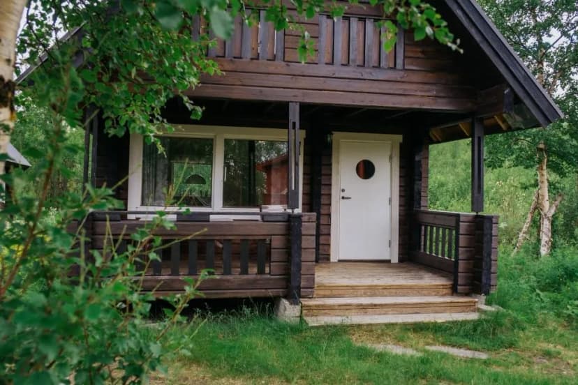 Dark wood cabin exterior with white door and porch surrounded by green foliage, Abisko Mountain Lodge
