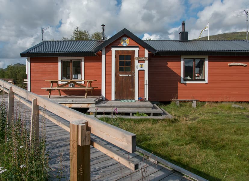 Red mountain hut with wooden walkway and picnic table at Abiskojaure under cloudy sky.
