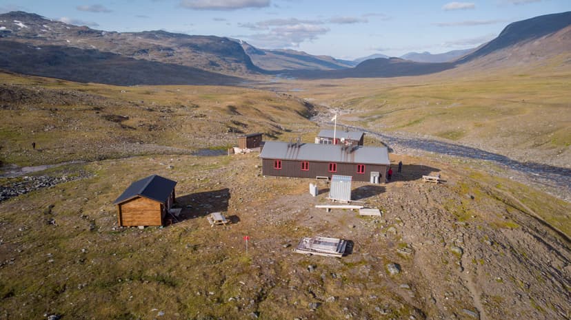 Mountain hut complex in rocky, grassy valley with surrounding barren mountains and stream.