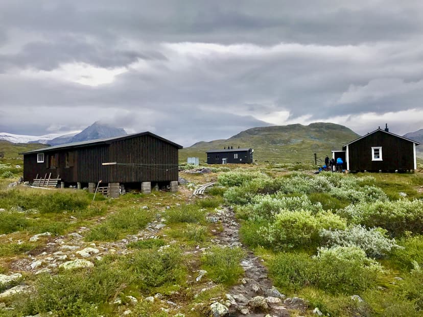 Dark wooden huts in a rocky, brush-covered mountain landscape under a cloudy sky, likely Singi Hut.