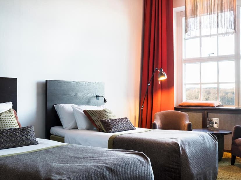 Twin beds in a hotel room with white walls, red curtains, and a view of trees outside the window.