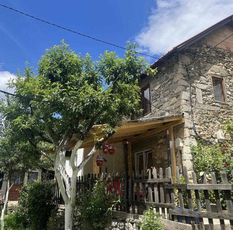 Stone guest house with wooden porch, white-painted tree, and picket fence under blue sky.