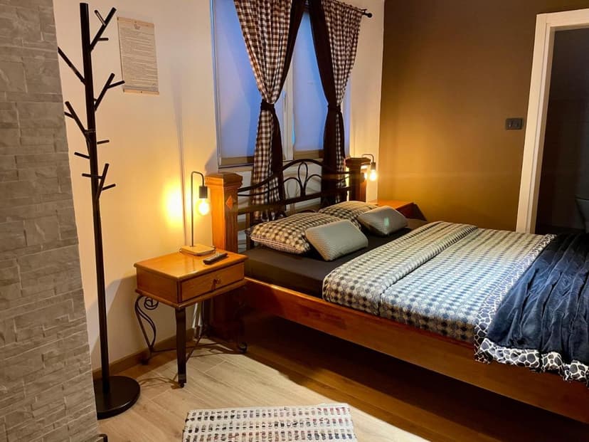 Cozy guest room with wooden bed, patterned bedding, and warm lighting near a window with checkered curtains.