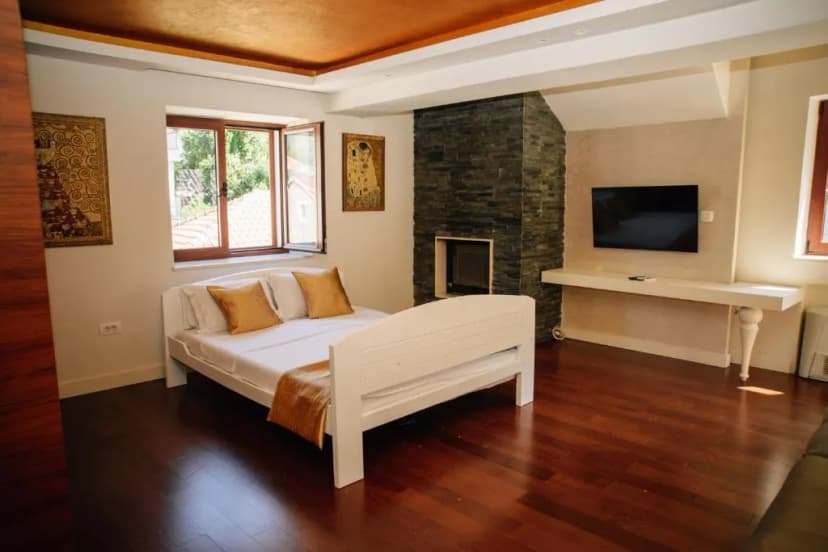 Deluxe room with white bed, stone fireplace, wall-mounted TV, and dark wood floor
