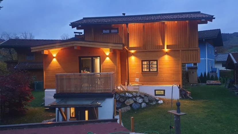 Wooden chalet exterior illuminated at dusk with a small balcony and green lawn in a hilly area.