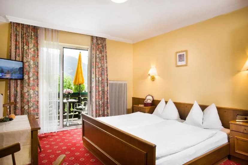 Hotel room with wooden bed, floral curtains, and balcony view of green mountains at Landhotel Steindlwirt.