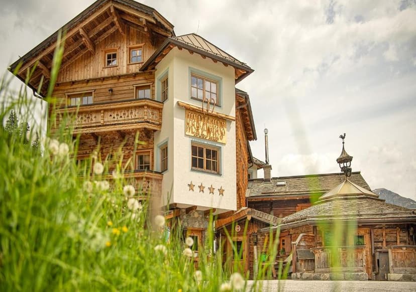 Alpine hotel with wooden balconies viewed through tall green grass and dandelions.