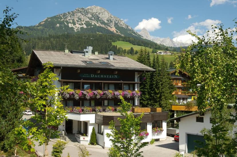 Hotel Dachstein building with flower boxes against a backdrop of green mountains and blue sky.