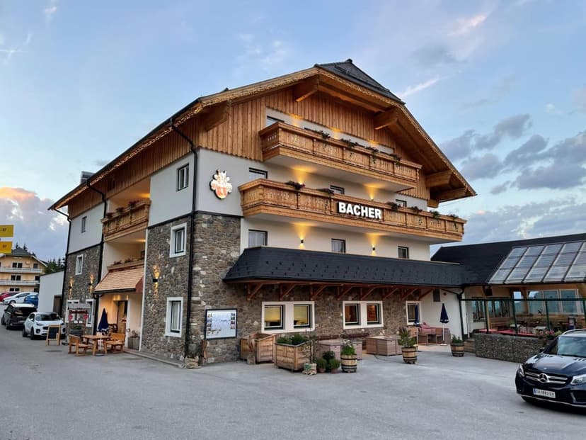 Gasthof Bacher building with stone facade, wooden balconies, and illuminated sign at dusk.