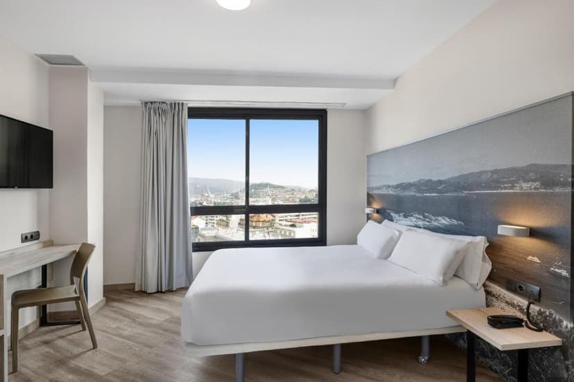 Hotel room with white bedding, desk, and city view from window in Vigo.