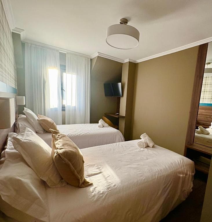 Twin beds with white linens in a hotel room with a window and wall-mounted television.