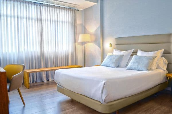 Hotel Rías Baixas bedroom with white bedding, light blue pillows, and sheer window curtains.