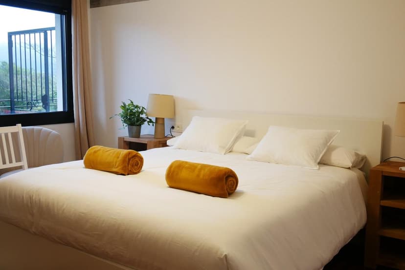 Bedroom with white bedding, two rolled yellow blankets, and a window view of greenery.