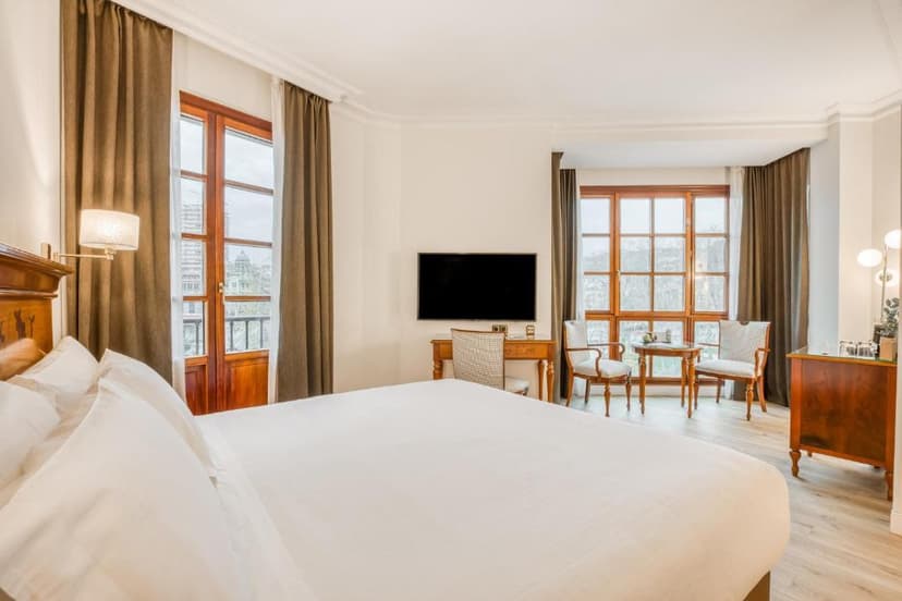 Hotel bedroom with large white bed, desk, seating area, and city view through wooden windows.