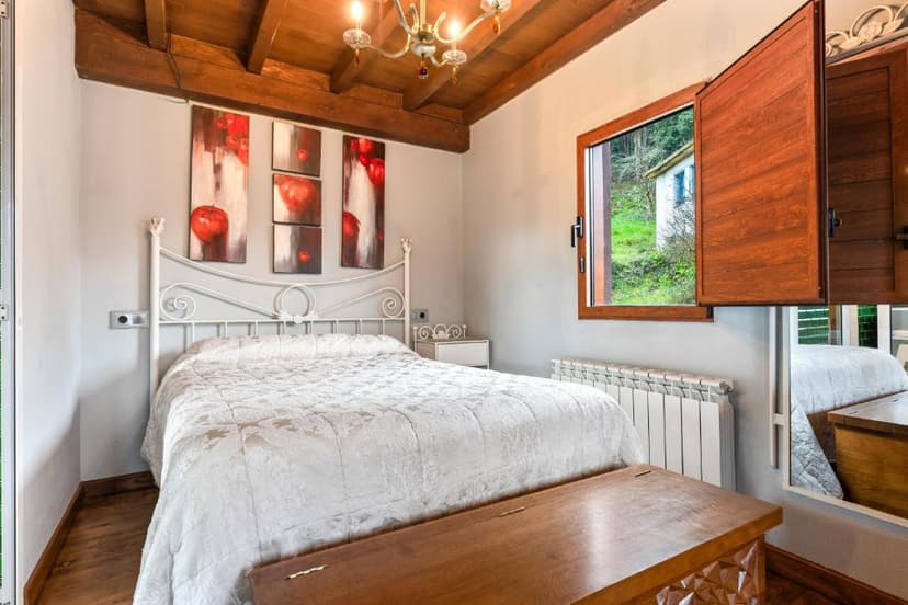 Bedroom with white metal bed, wooden ceiling beams, and view of green hillside outside.