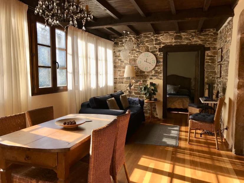 Rustic living and dining area with stone walls, wood beams, and bright sunlight through sheer curtains