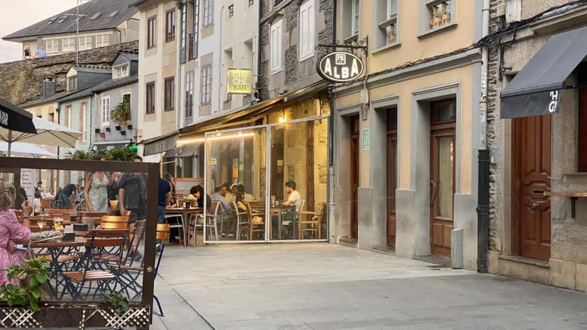 Outdoor dining patio next to Hostal Alba in Lugo with people seated at tables on a street.