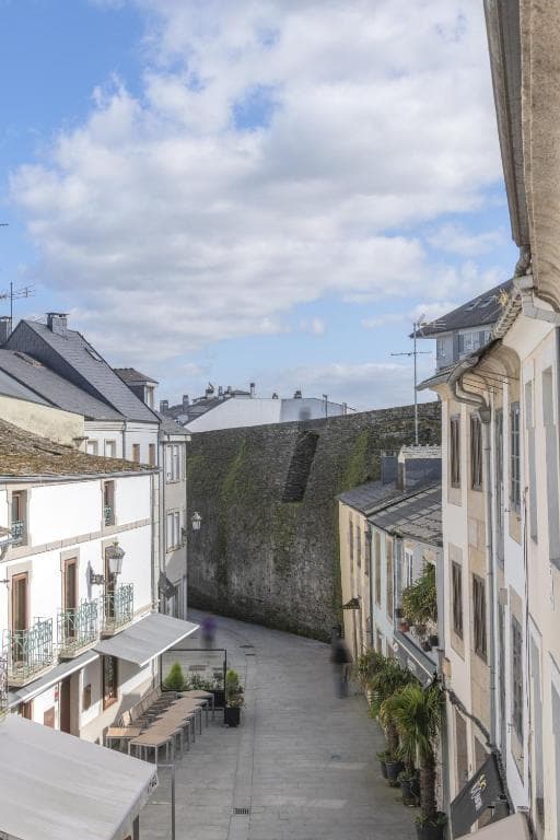 Street view in Lugo, Spain, showing white buildings and a tall mossy stone wall under a cloudy sky.