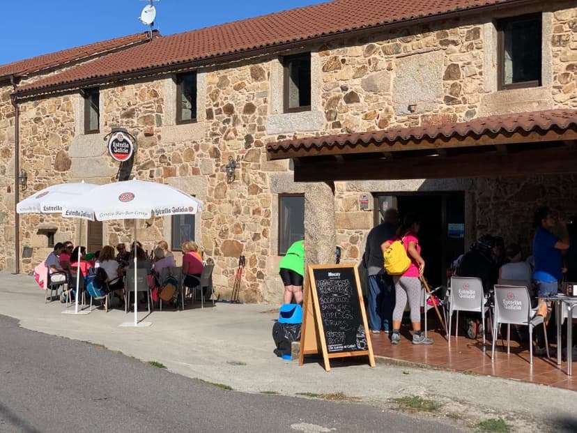 Albergue Ponte Olveira with outdoor seating under an umbrella, featuring stone building and chalkboard menu.