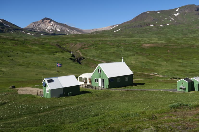 Green huts at Húsavíkurskáli with Icelandic flag, set against grassy hills and mountains.