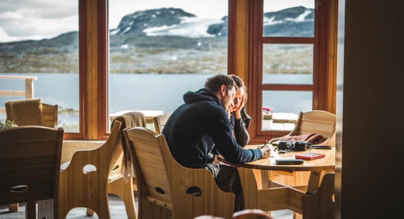 Couple at wooden table inside cabin looking out at snowy mountains and lake near Finsehytta.