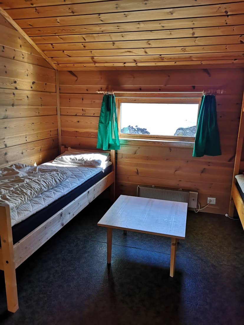 Simple wooden cabin interior with beds, table, and window view of snowy rocks at Geiterygghytta.
