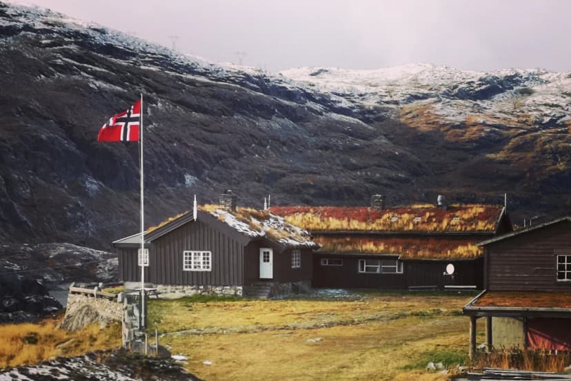 Steinbergdalshytta cabin with grass roof, Norwegian flag, and snow-dusted mountains.