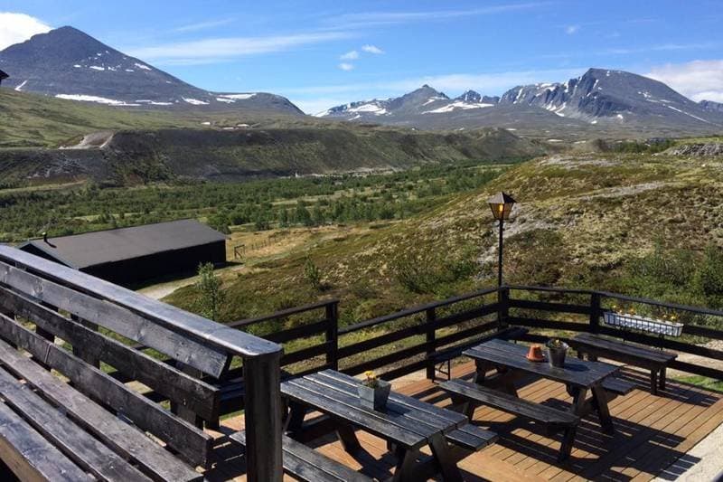 Wooden deck with picnic tables overlooking green valley and snow-capped mountains.