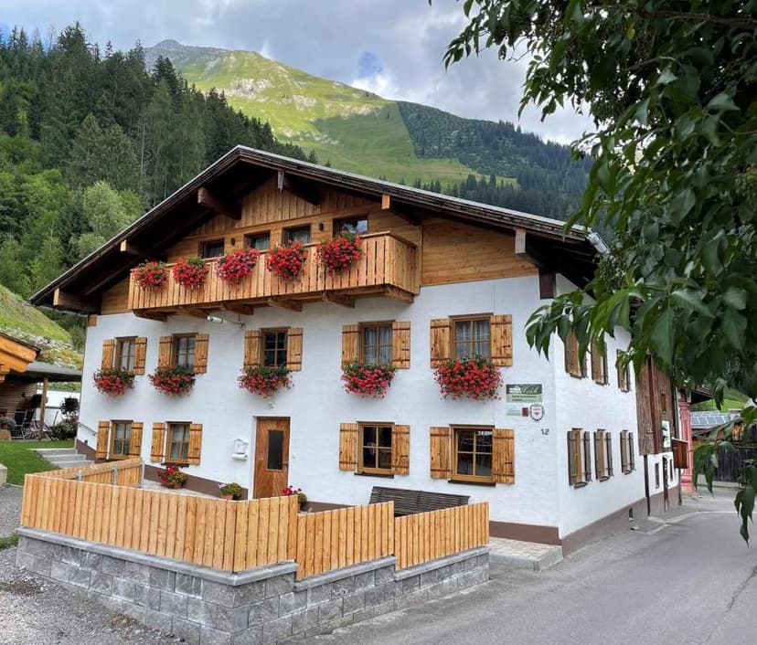 Traditional Alpine house with wooden balconies and red flowers against a forested mountain backdrop.