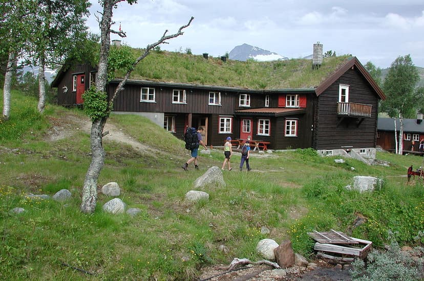 Hikers approach a dark wooden cabin with a grass roof, snow-capped mountain in background.