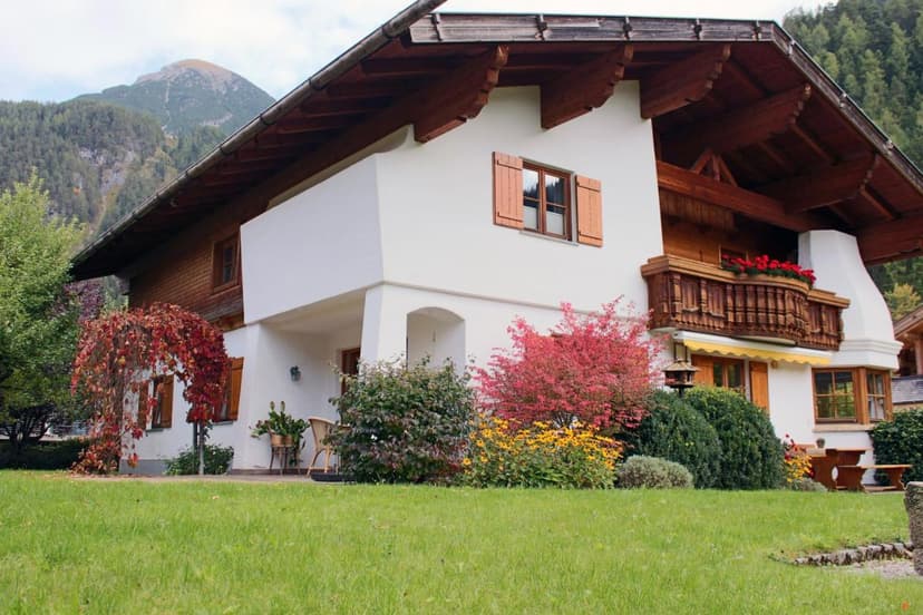 White alpine vacation home with wooden balcony, colorful autumn shrubs, and mountain backdrop.