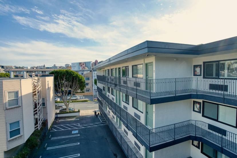Two-story motel building with exterior corridors and black railings, parking lot, and city background.