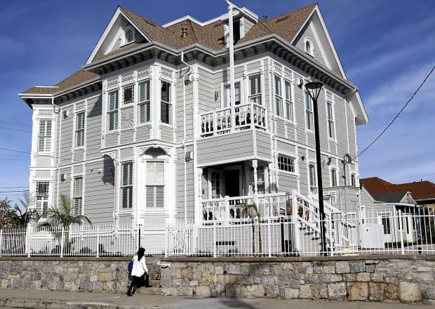 Victorian-style inn with gray siding and white trim, set above a stone retaining wall.