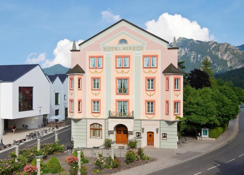 Hotel Hirsch building with pink facade, set against lush green mountains under a blue sky.