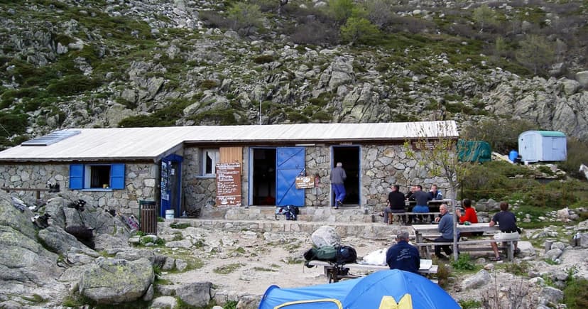 Refuge d'Usciolu stone mountain hut with hikers resting outside near tents and rocky terrain.