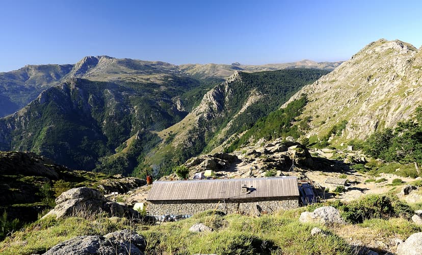 Refuge d'Usciolu stone mountain hut with wooden roof surrounded by steep, forested mountains.